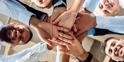 Gruppe von lachenden Menschen steht im Kreis und legt ihre Hände übereinander in die Mitte.